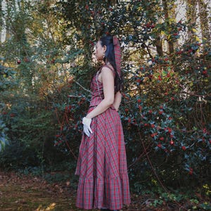 May include: A red plaid dress with a matching headscarf and white gloves. The dress has a sleeveless top and a long skirt. The person is wearing black platform boots. The background is a forest with green trees and red berries.
