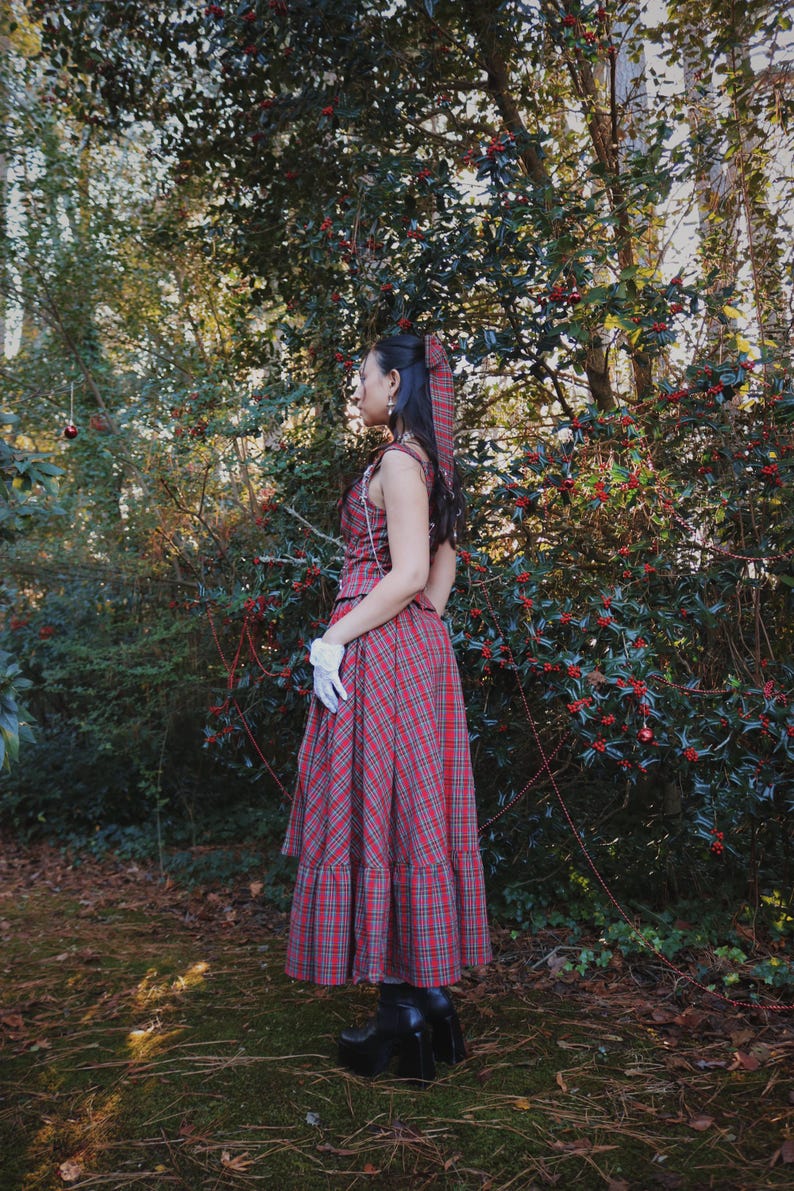 May include: A red plaid dress with a matching headscarf and white gloves. The dress has a sleeveless top and a long skirt. The person is wearing black platform boots. The background is a forest with green trees and red berries.