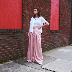 May include: A woman wearing a white crop top, a white cardigan, and wide-legged pink satin pants. She is standing in front of a brick wall with red painted panels.