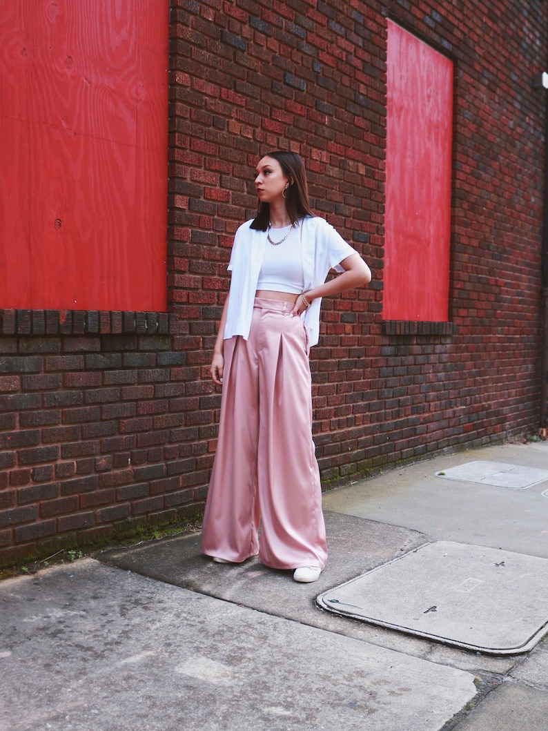 May include: A woman wearing a white crop top, a white cardigan, and wide-legged pink satin pants. She is standing in front of a brick wall with red painted panels.