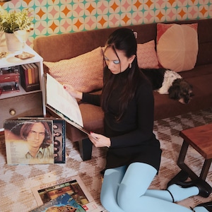 May include: A woman in a black dress and light blue tights sits on the floor, surrounded by vinyl records. The room has a retro aesthetic with a patterned wallpaper and a brown sofa. A dog rests on the sofa.