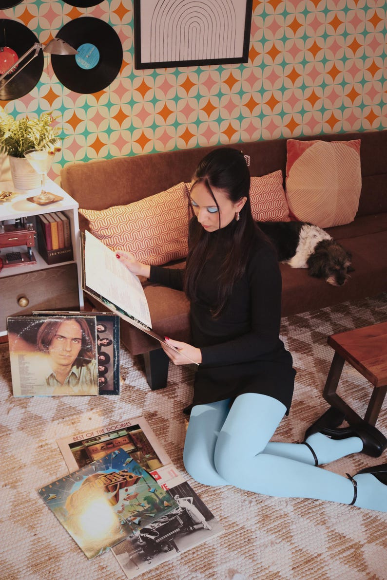 May include: A woman in a black dress and light blue tights sits on the floor, surrounded by vinyl records. The room has a retro aesthetic with a patterned wallpaper and a brown sofa. A dog rests on the sofa.
