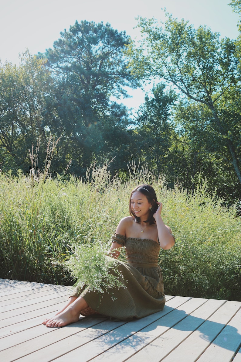Op de afbeelding: Een vrouw in een groene, off-shoulder jurk zit op een houten dek en houdt een boeket witte bloemen vast. Ze wordt omringd door groen gebladerte en bomen.