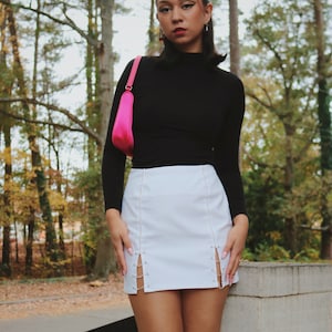 May include: A woman wearing a black turtleneck shirt and a white mini skirt with silver chain detailing. She is standing in a wooded area and has a pink handbag slung over her shoulder.