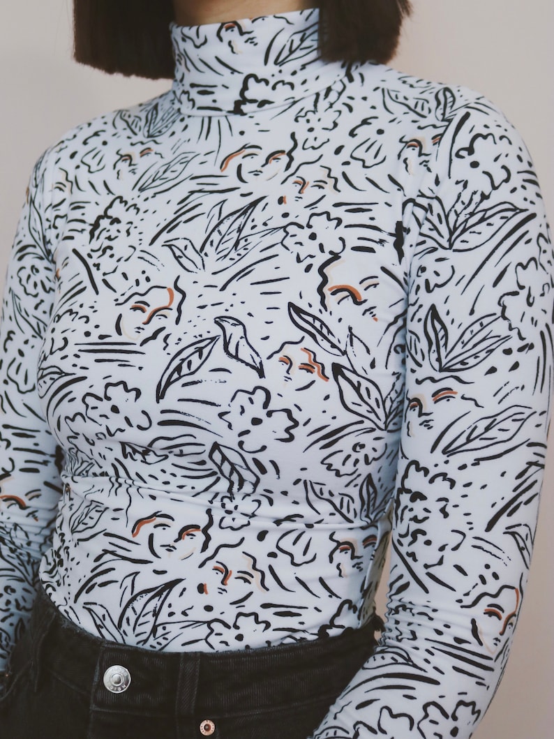 May include: A long-sleeved, light blue turtleneck shirt with a black and brown abstract print. The print features stylized leaves, flowers, and swirls.