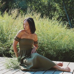 Op de afbeelding: Een vrouw in een groene, off-shoulder jurk zit op een houten dek met een boeket witte bloemen in haar schoot. Ze is omringd door hoog groen gras en bomen.
