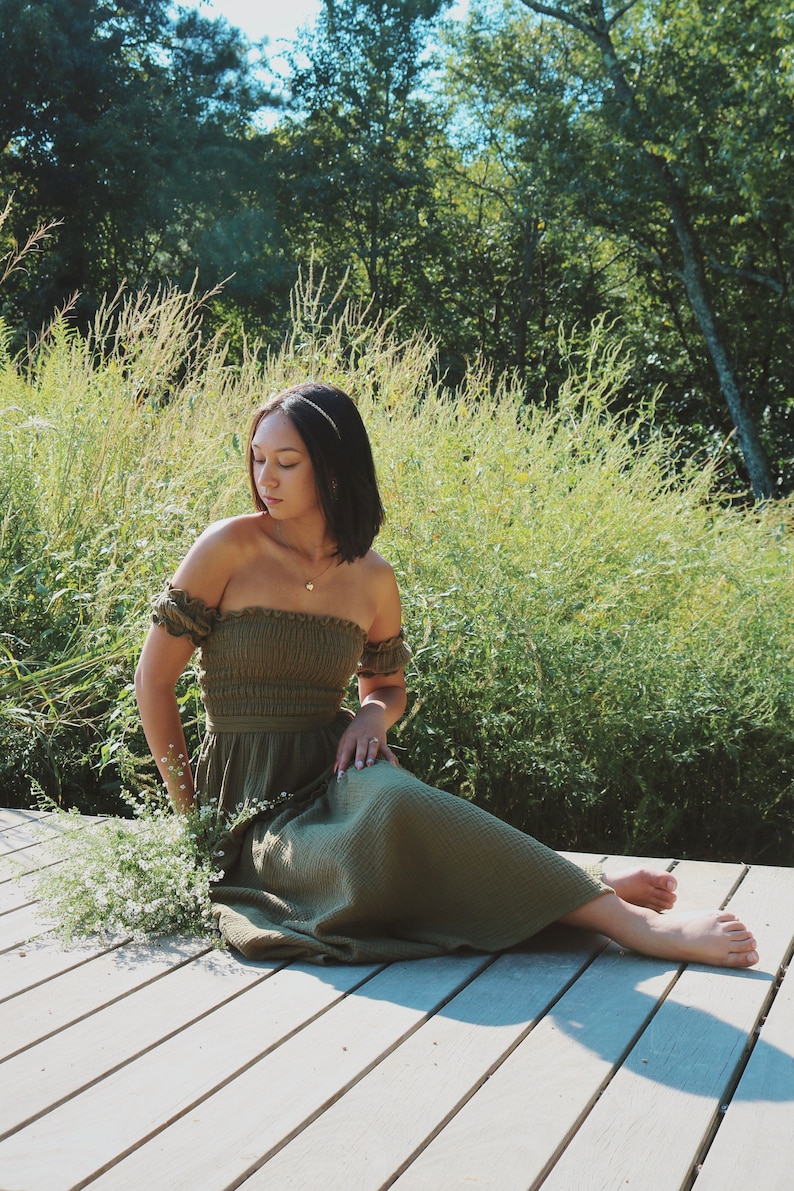 Op de afbeelding: Een vrouw in een groene, off-shoulder jurk zit op een houten dek met een boeket witte bloemen in haar schoot. Ze is omringd door hoog groen gras en bomen.