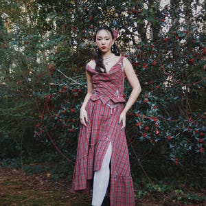 May include: A red plaid dress with a matching top, featuring a high slit and a layered skirt. The outfit is paired with black platform boots and white tights. The model is standing in front of a green bush with red berries.