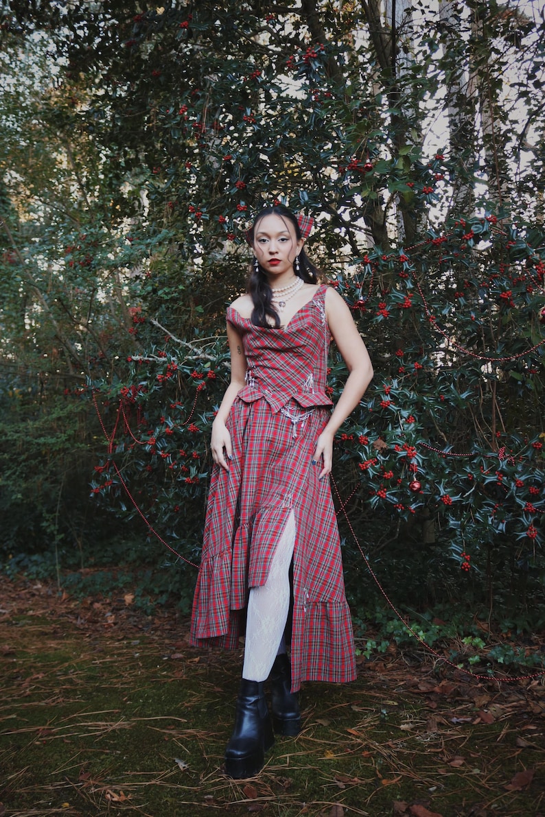 May include: A red plaid dress with a matching top, featuring a high slit and a layered skirt. The outfit is paired with black platform boots and white tights. The model is standing in front of a green bush with red berries.