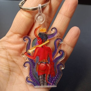 May include: A clear acrylic keychain featuring a red demon-like figure with a crown, holding a sword, and surrounded by purple tentacles. The keychain has a silver keyring attached.