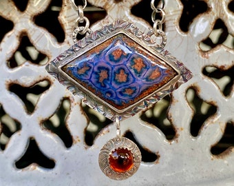 Può includere: Una collana con catena d'argento con un ciondolo a forma di diamante con un designo in smalto blu e arancione. Il ciondolo è attaccato a un ciondolo rotondo più piccolo con una pietra arancione.