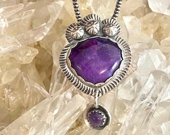 May include: A silver necklace with a purple stone pendant. The pendant is surrounded by three silver flowers and has a smaller purple stone dangling from the bottom.