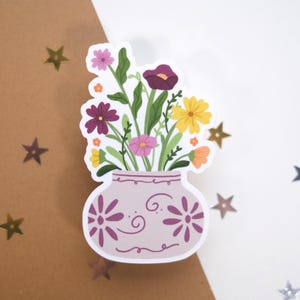 May include: A sticker featuring a vase filled with colorful flowers. The vase is light purple with floral designs. The flowers include purple, pink, yellow, and orange blooms with green leaves. The sticker is on a white and brown background with star-shaped confetti.