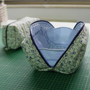 May include: Two quilted cosmetic bags. The open bag features a blue and white floral pattern on a light green fabric, with a blue zip and a blue and white gingham lining. The second bag is closed.