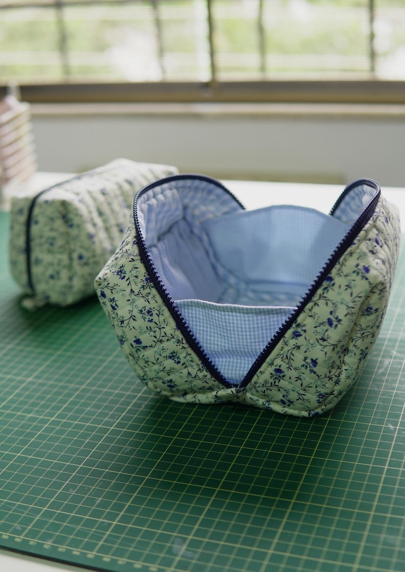 May include: Two quilted cosmetic bags. The open bag features a blue and white floral pattern on a light green fabric, with a blue zip and a blue and white gingham lining. The second bag is closed.