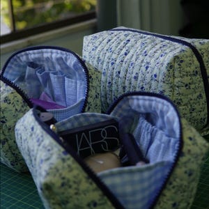 May include: Three quilted cosmetic bags featuring a blue floral pattern on a light background. The bags have dark blue zips and a light blue and white gingham interior lining with elasticated pockets containing various makeup items.