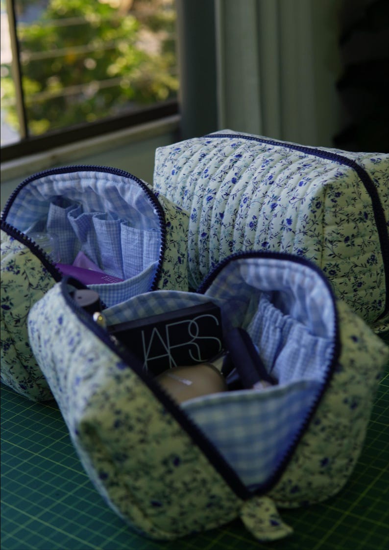 May include: Three quilted cosmetic bags featuring a blue floral pattern on a light background. The bags have dark blue zips and a light blue and white gingham interior lining with elasticated pockets containing various makeup items.
