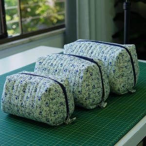 May include: Three quilted cosmetic bags in different sizes, displayed on a green surface. The bags have a white background with a blue floral design and a navy blue zip. The bags are made of a soft, quilted fabric.