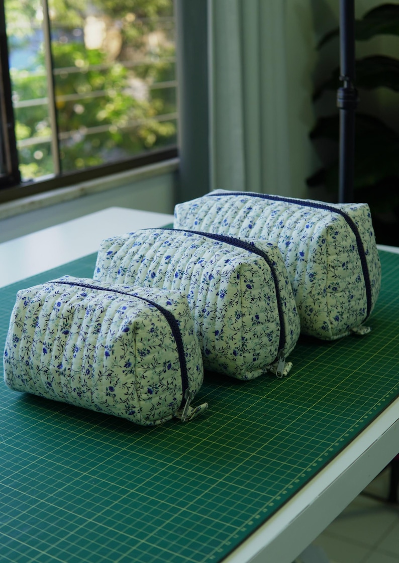 May include: Three quilted cosmetic bags in different sizes, displayed on a green surface. The bags have a white background with a blue floral design and a navy blue zip. The bags are made of a soft, quilted fabric.