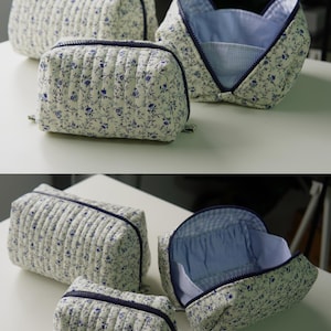 May include: A set of three quilted pouches featuring a white fabric with a blue floral pattern and dark blue zips. The pouches have a rectangular shape with vertical stitching and a light blue gingham interior lining.
