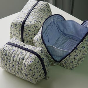 May include: Three quilted cosmetic bags in different sizes. They have a cream floral pattern with blue details and dark blue zippers. One bag is open, showing a blue and white gingham lining.