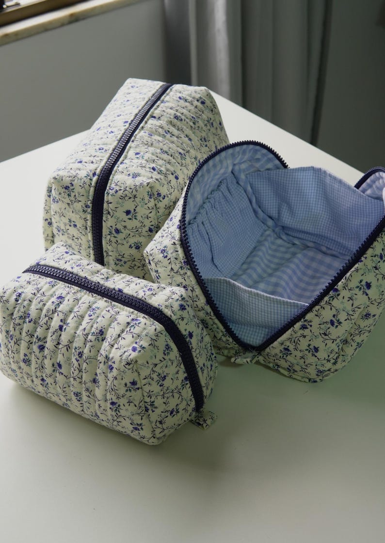 May include: Three quilted cosmetic bags in different sizes. They have a cream floral pattern with blue details and dark blue zippers. One bag is open, showing a blue and white gingham lining.