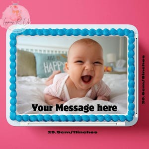 May include: An edible cake topper featuring a photo of a smiling baby. The topper has a blue border and the text "Your Message here". The topper measures 29.5cm wide and 20.6cm tall.