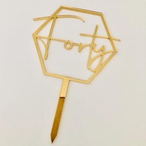 May include: Gold-colored cake topper with a geometric hexagon shape and the word "forty" written inside. The topper is on a thin, gold-colored stick.