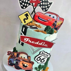 May include: A birthday cake decorated with characters from the Cars movie, including Lightning McQueen and Mater. The cake is light blue with a Route 66 sign, checkered flags, and the name "Freddie".