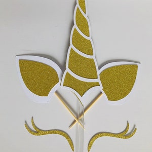May include: Gold glitter unicorn cake topper with white outlines. The topper includes a horn, ears, and eyelashes. The topper is attached to two wooden skewers.
