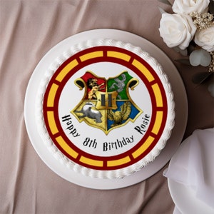 May include: A round white cake with a decorative border and the text "Happy 8th Birthday Rosie". The cake features a colorful emblem with a crest including a badger, eagle, lion, and snake. The cake is on a white plate.