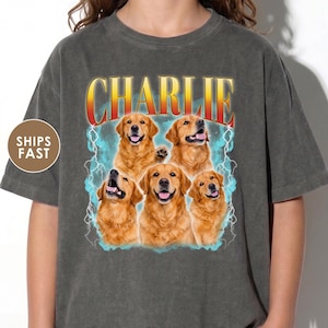 May include: A charcoal gray short-sleeve t-shirt featuring a graphic print of five golden retriever dogs. Above the dogs, the name CHARLIE is printed in a bold, yellow-to-red gradient font. The background of the graphic includes light blue lightning.
