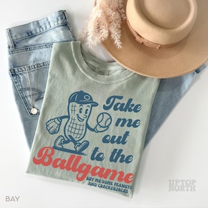 May include: A light green t-shirt with a vintage-style illustration of a peanut wearing a baseball cap and holding a baseball. The text "Take me out to the Ballgame" is printed in blue and red on the shirt. The text "Buy me some peanuts and crackerjacks" is printed in smaller blue text below the illustration.