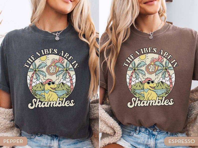 May include: Two t-shirts, one pepper and one espresso, featuring a circular graphic with a cartoon dinosaur wearing sunglasses. The graphic includes the text "THE VIBES ARE IN Shambles" with palm trees, a sun, and a meteor.