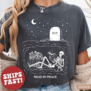 May include: A black t-shirt with a white graphic of a skeleton reading a book in a graveyard. The text "Read in Peace" is printed below the graphic.