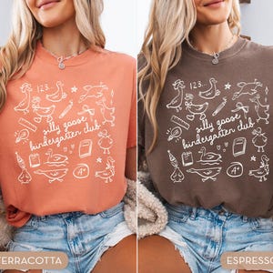 K&ouml;nnte beinhalten: Zwei T-Shirts, eines in Terrakotta und eines in Espresso, mit einer wei&szlig;en Grafik. Die Grafik enth&auml;lt den Text "silly goose kindergarten club" mit Illustrationen von G&auml;nsen, Schulmaterial und den Zahlen "123".