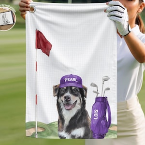 Op de afbeelding: Een witte golfhanddoek met een gedrukte illustratie van een hond die een paarse pet draagt met de naam PEARL, naast een paarse golf-tas met het label LOIS, een rode vlag aan een paal en een green op een witte, getextureerde achtergrond.