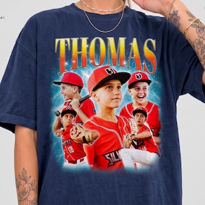 May include: A navy blue t-shirt featuring a graphic print with the name THOMAS in large yellow and orange letters. The design includes multiple images of a baseball player in a red uniform and cap, set against a blue background.