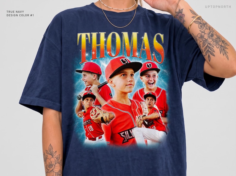 May include: A navy blue t-shirt featuring a graphic print with the name THOMAS in large yellow and orange letters. The design includes multiple images of a baseball player in a red uniform and cap, set against a blue background.