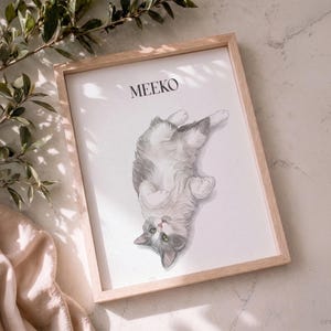 Op de afbeelding: Een rechthoekige lijst van licht hout met een witte print van een grijs-witte kat die op de rug ligt met de pootjes ingetrokken. De naam MEEKO staat in zwarte hoofdletters boven de illustratie van de kat op de witte achtergrond.