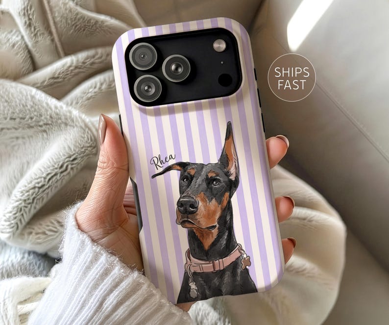 May include: A smartphone case featuring a vertical lavender and white striped pattern with a central illustration of a black and tan Doberman dog wearing a light pink collar. The name Rhea is printed in a cursive script next to the dog's head.