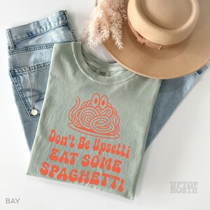 May include: A sage green t-shirt with the text "Don't Be Upsettti EAT SOME SPAGHETTI" in orange. The shirt is paired with light blue jeans, a tan hat, and a gold necklace. Dried flowers are also in the image.