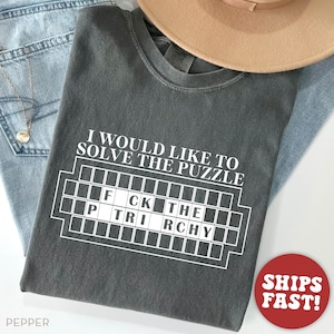 May include: A dark gray t-shirt with the text "I would like to solve the puzzle" and a white grid with the words "FCK THE TRCHY" inside. The shirt is made of a soft, comfortable material.