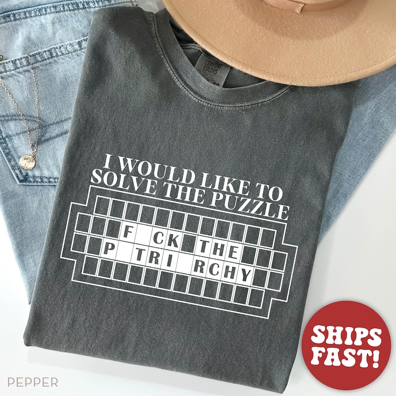 May include: A dark gray t-shirt with the text "I would like to solve the puzzle" and a white grid with the words "FCK THE TRCHY" inside. The shirt is made of a soft, comfortable material.