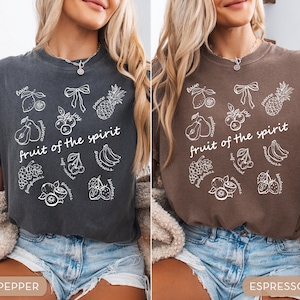 May include: Two t-shirts, one pepper-colored and one espresso-colored, each featuring a white graphic. The graphic includes the text "fruit of the spirit" and illustrations of various fruits and words like "love" and "peace".