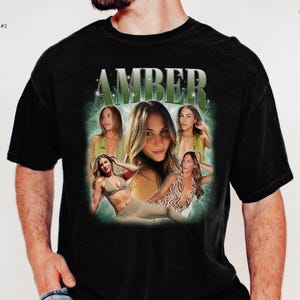 May include: A black short-sleeve t-shirt featuring a graphic print with the name AMBER in large green letters at the top. The design includes a collage of four photographs of a woman in various poses, set against a glowing green background.