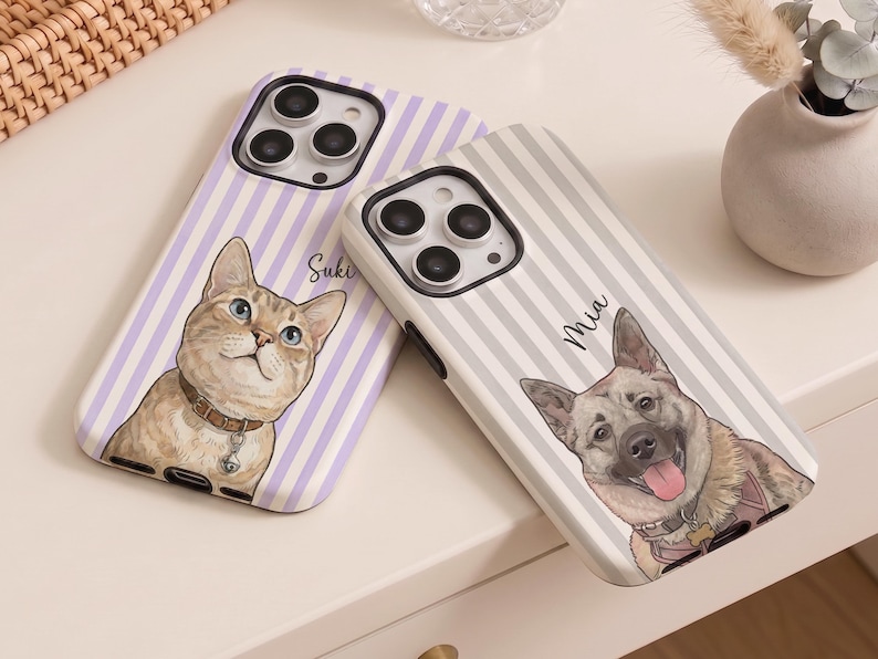 May include: Two smartphone cases featuring custom pet portraits. The left case has a light purple striped pattern with a cat illustration and the name Suki. The right case has a grey striped pattern with a dog illustration and the name Mia.