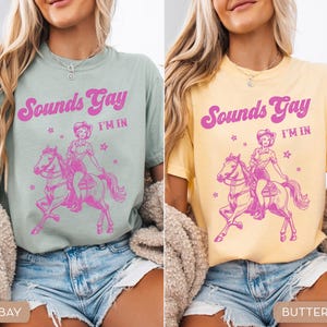 K&ouml;nnte beinhalten: Zwei Frauen tragen T-Shirts mit dem Text "Sounds Gay I'm In" in rosa Buchstaben. Die Shirts zeigen eine rosa Illustration einer Frau, die auf einem Pferd reitet. Die Frau links tr&auml;gt ein hellgr&uuml;nes Shirt und die Frau rechts ein gelbes Shirt.