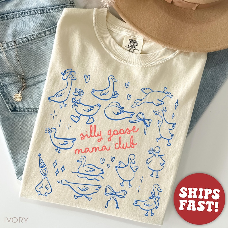 May include: Ivory-colored t-shirt with a blue duck illustration and the text "silly goose mama club" in red. The design features various duck drawings, hearts, and bows. The shirt is folded on top of blue jeans.