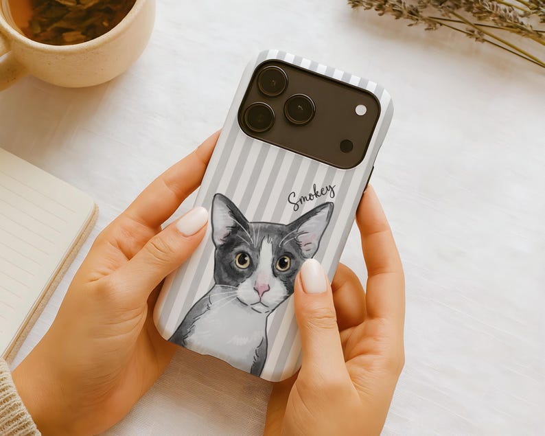 May include: A smartphone case featuring a light gray and white vertical striped pattern. The case displays a detailed illustration of a gray and white cat with large eyes and the name Smokey written in cursive script above the cat's head.